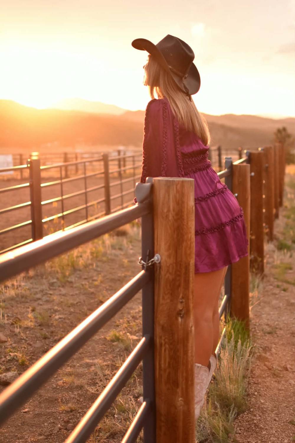 A woman in a hat leaning on a pipe fence at sunset, framed in oak.