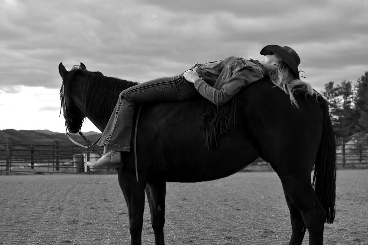 A woman lying back on a black horse, hat over her face, in black and white.